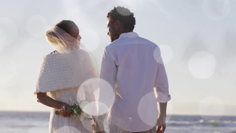 Animation-Von-Lichtpunkten-über-Afroamerikanischer-Braut-Und-Bräutigam,-Die-Bei-Einer-Hochzeit-Am-Strand-Händchen-Halten