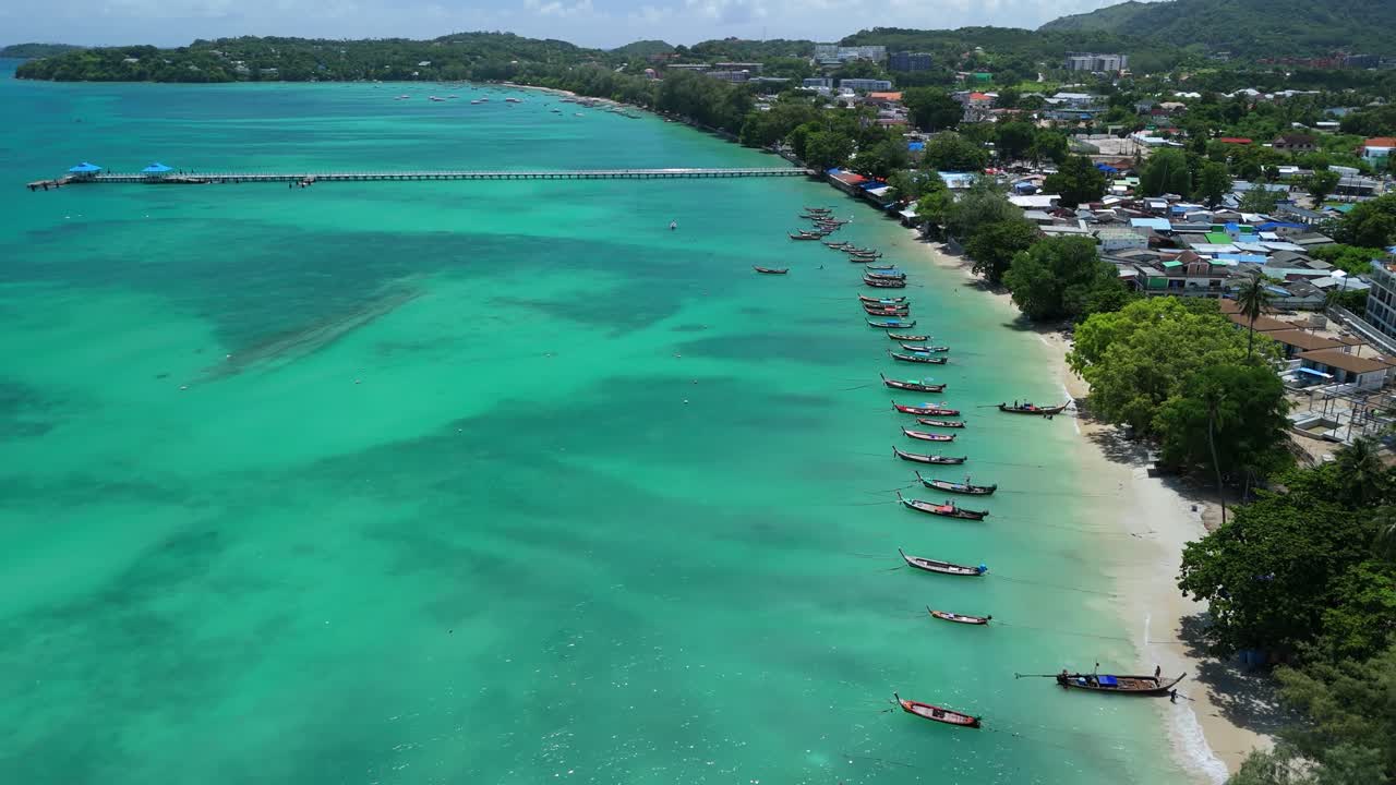 Drone pushes back above moored longtail boats along turquoise Rawai Beach, Phuket