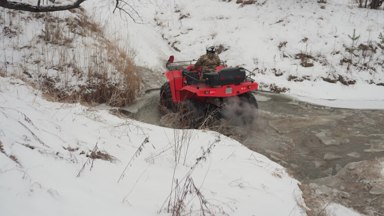 ATV rosso che attraversa un ruscello ghiacciato, il pilota naviga in un torrente ghiacciato e poco profondo con acqua che schizza e cumuli di neve. Una ripresa ravvicinata cattura gli schizzi di fango, il pilota con il casco si china sull'acceleratore. Atmosfera off-road selvaggia.