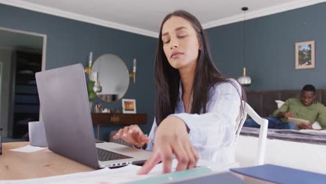 Video-of-biracial-woman-using-laptop-and-reading-notes,-working-in-bedroom,-partner-in-background