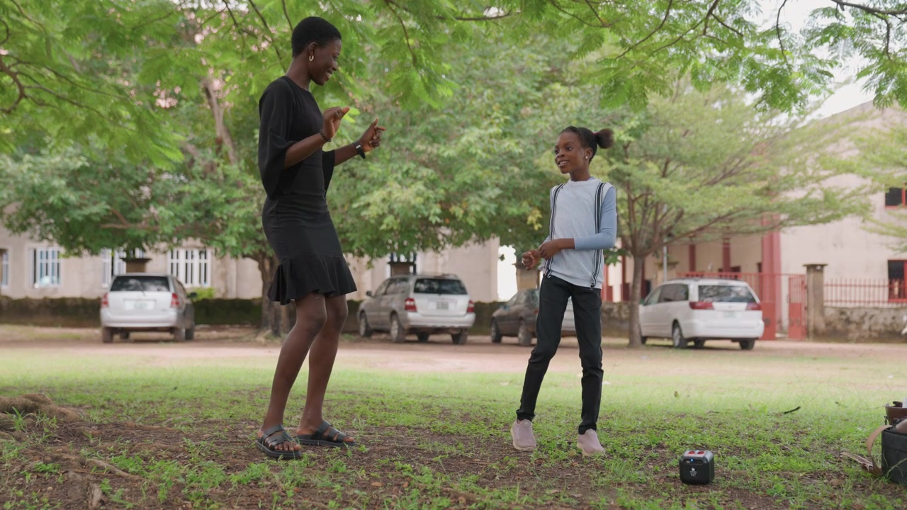 Black mother and daughter under tree, sharing tender guidance beside small speaker on grass, daughter in school uniform with backpack, mother in black dress offering playful instruction, suburban