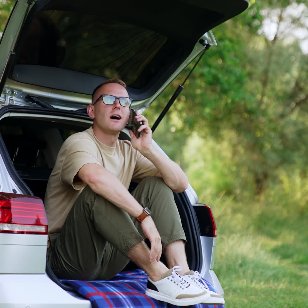 Relaxed Caucasian man holding smartphone. Man starts to talk on the phone sitting in the car trunk. Forest at backdrop