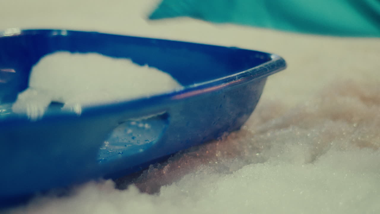 아이는 차가운 추위를 피하기 위해 재으로 인 손으로 파란 플라스틱 용기에 얼음을 모으고 눈에서 놀고 있습니다.