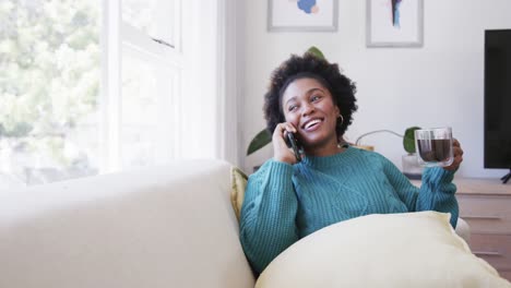 Fröhliche-Afroamerikanische-Frau-Entspannt-Sich-Auf-Der-Couch,-Spricht-Auf-Dem-Smartphone-Und-Trinkt-Tee,-Zeitlupe