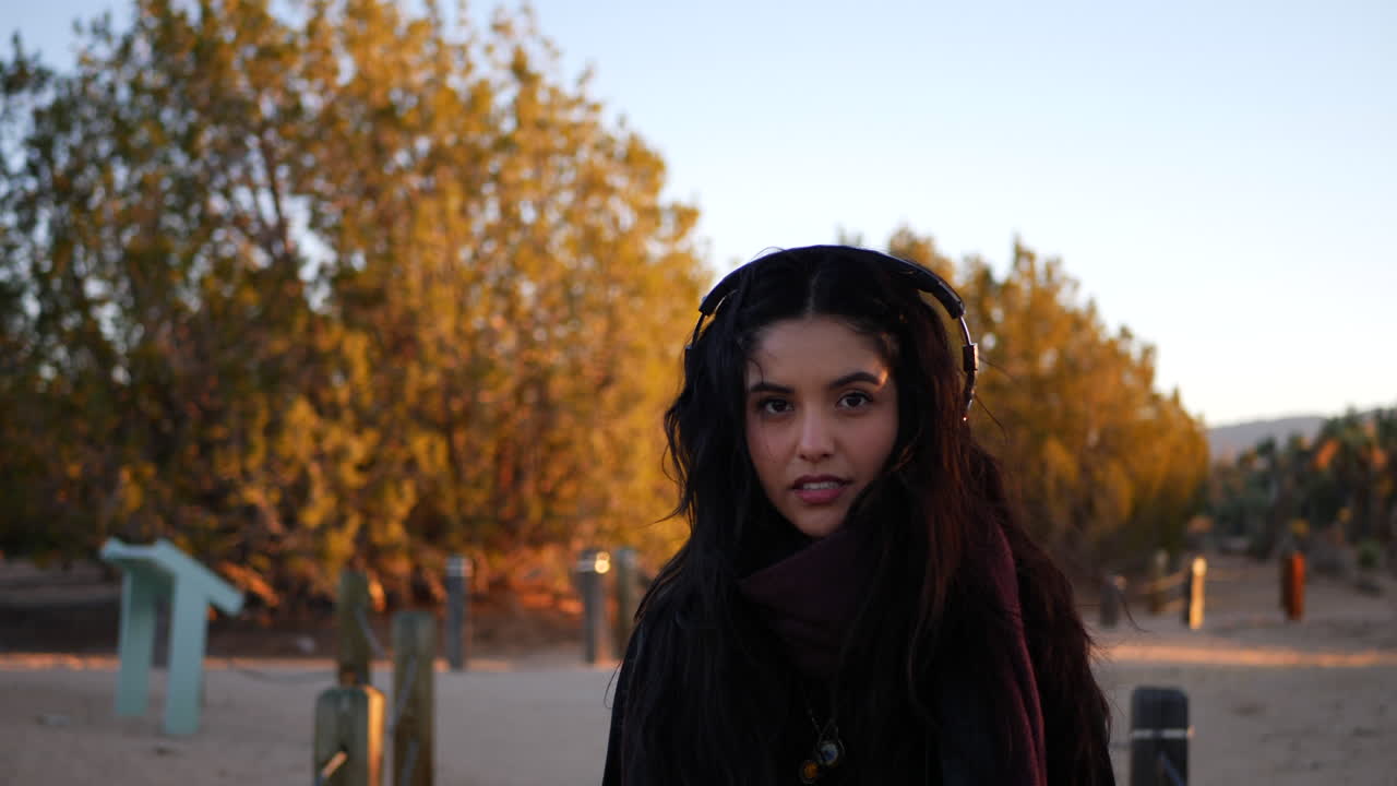 una hermosa mujer caminando en cámara lenta al atardecer de la hora dorada escuchando música con auriculares mientras piensa en su vida