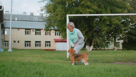canine specialist exercising on grassy field near goal post, smiling while being distracted by playful puppy wanting attention, surrounded by trees and building background showing outdoor training