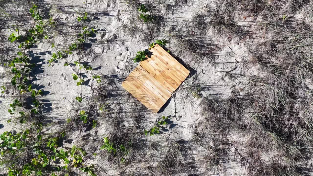 A drone camera smoothly descends toward a square wooden landing pad set on coastal sand dunes, surrounded by sparse vegetation in bright daylight