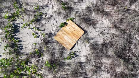 a drone camera smoothly descends toward a square wooden landing pad set on coastal sand dunes, surrounded by sparse vegetation in bright daylight