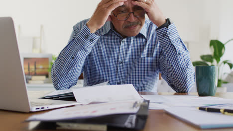 Video-of-stressed-senior-biracial-man-sitting-at-desk-looking-at-paperwork-and-using-laptop-at-home