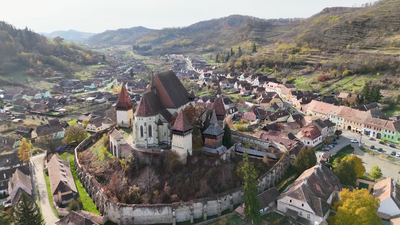 As the drone moves closer, the vibrant autumn colors of the landscape and the charming village below come into focus, showcasing the perfect harmony of history, architecture, and nature.