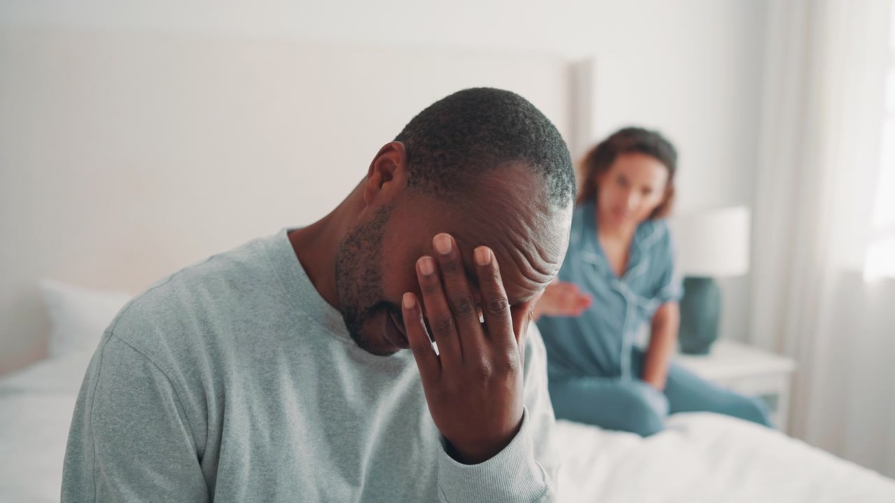 Uomo stressato seduto sul letto mentre il partner litiga sullo sfondo