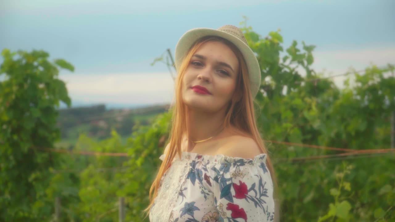 impresionantes imágenes en hd de una chica caucásica con un sombrero de punto y lápiz labial rojo de pie en un viñedo, iluminado por la luz del sol, mirando y sonriendo a la cámara