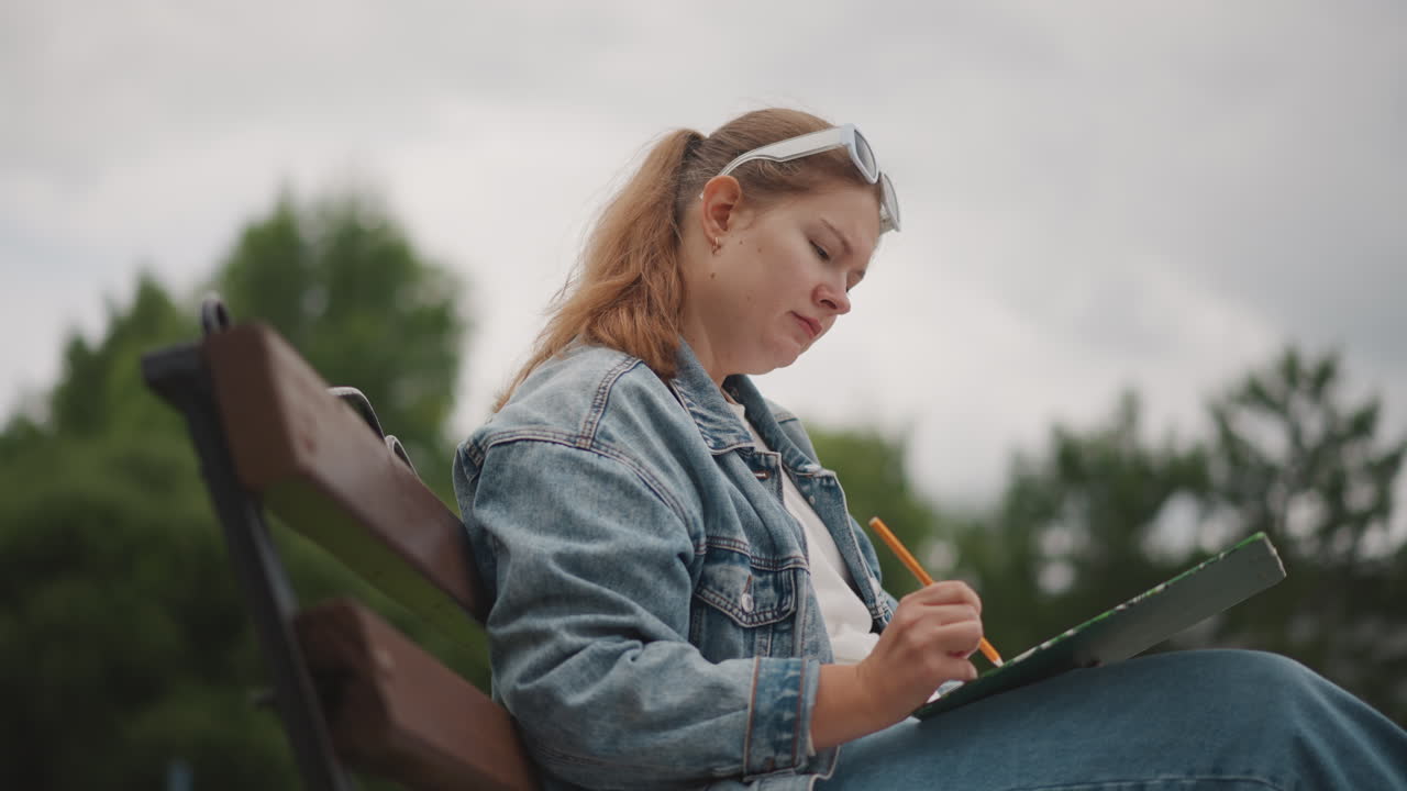 White woman sketching on bench, denim jacket and headband, pencil in hand, journal resting on lap, overcast sky, soft greenery and trees in background, intimate profile shot with calm reflective mood