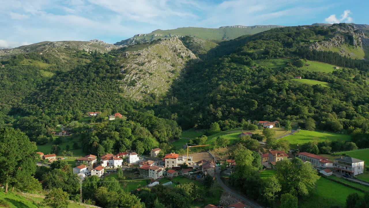 vuelo en un pueblo rural con su entorno agrícola y ganadero, sus verdes prados en verano y un fondo de bosques de robles y hayas con montañas de piedra caliza en cantabria, españa