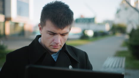 data analyst wearing black suit with silver buttons outdoors intensely focused on task, blurred background includes parked cars, green grass, pavement, and bench structures