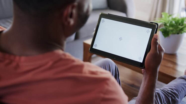 African American man noticing crosshair markers on tablet, adjusting angle in orange shirt on sofa