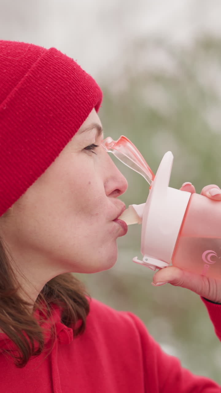 mom on maternity drinking water outdoors wearing red hoodie and beanie, holding pink bottle in serene natural setting with blurred background of greenery and light snow