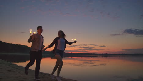 young happy multiethnic couple holding burning sparkling candles and running by the sea during sunset. slow motion shot