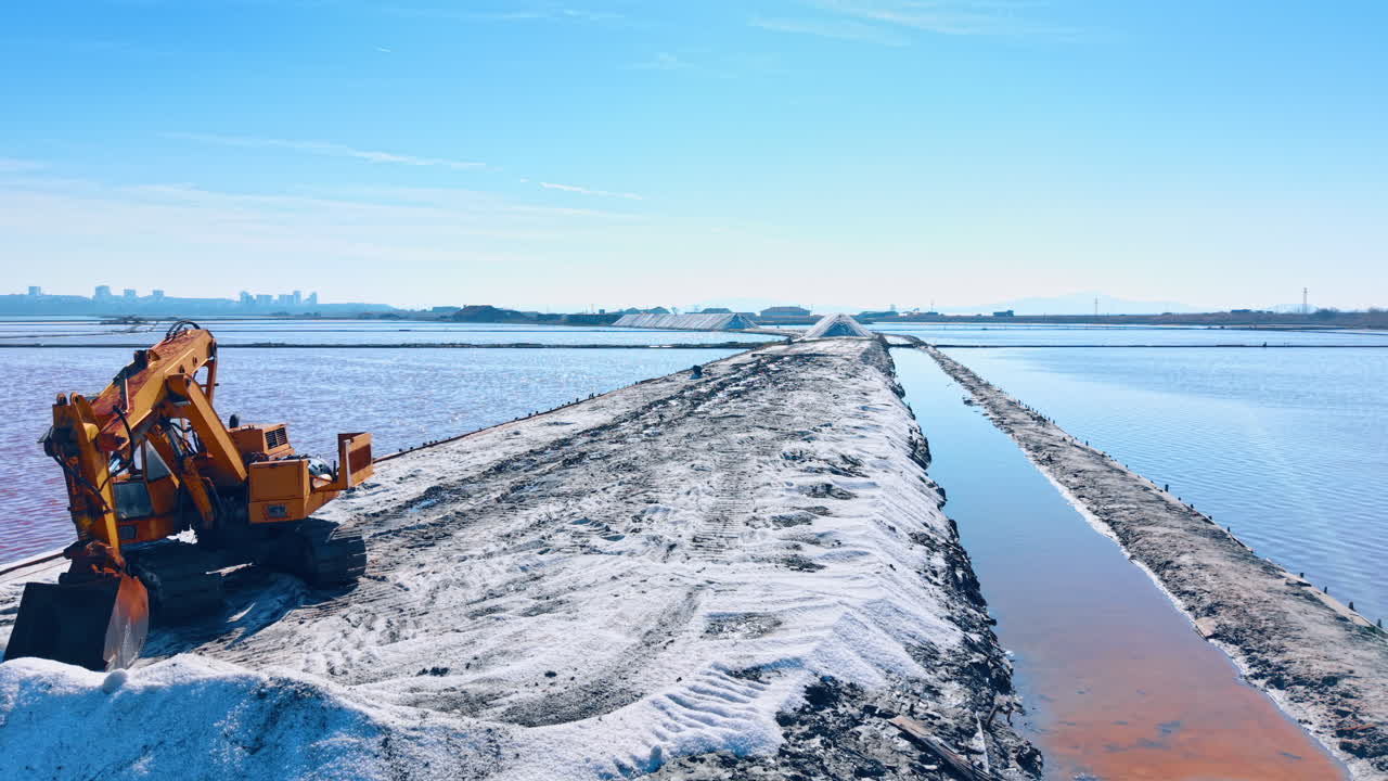 Escavatore giallo su un argine salino tra vasche d'acqua rosa. Scena industriale con un escavatore parcheggiato su un sentiero di sale bianco in una salina con lo skyline della città.