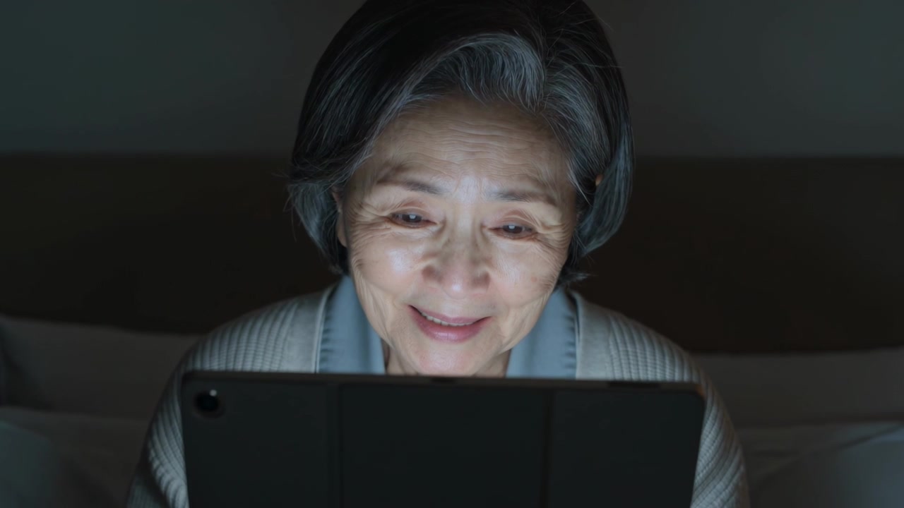 Reacting to tablet light, senior Asian woman holding tablet in bed and smiling, wearing cardigan