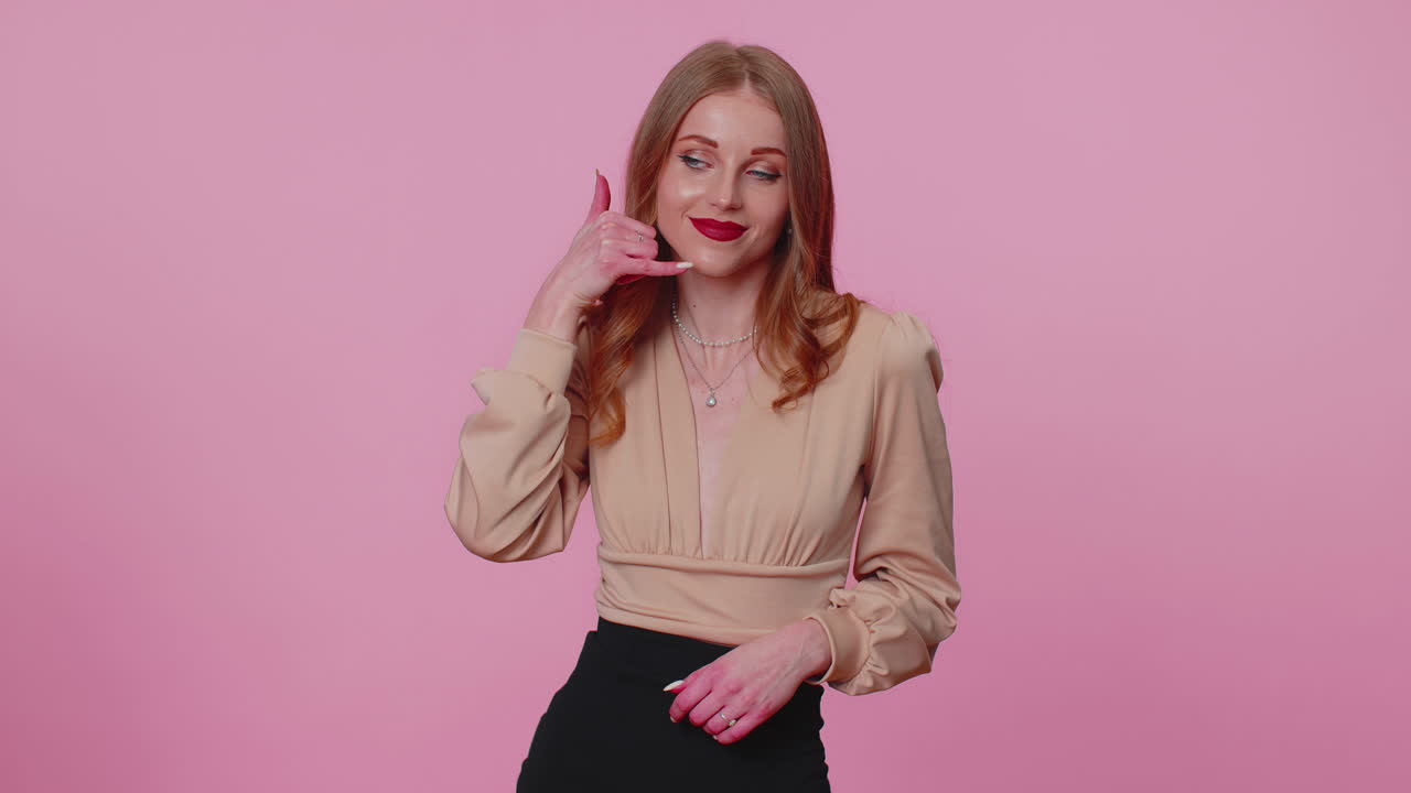 베이지 블라우스를 입은 유쾌한 소녀가 카메라를 바라보며 전화 제스처를 하고 있습니다.