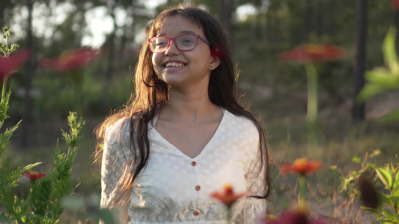 retrato de una niña con gafas vestida de blanco sonriendo en un campo de flores de zinnia, durante la puesta de sol