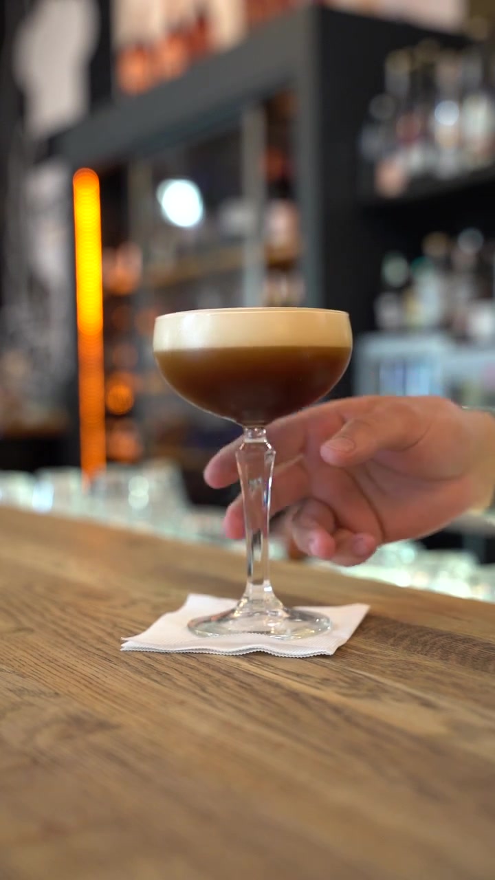 Un elegante espresso martini presentato in un calice con uno strato di schiuma cremosa, tenuto da una mano su un bancone da bar in legno. Lo sfondo è un ambiente da bar con illuminazione soffusa e scaffali con bottiglie.
