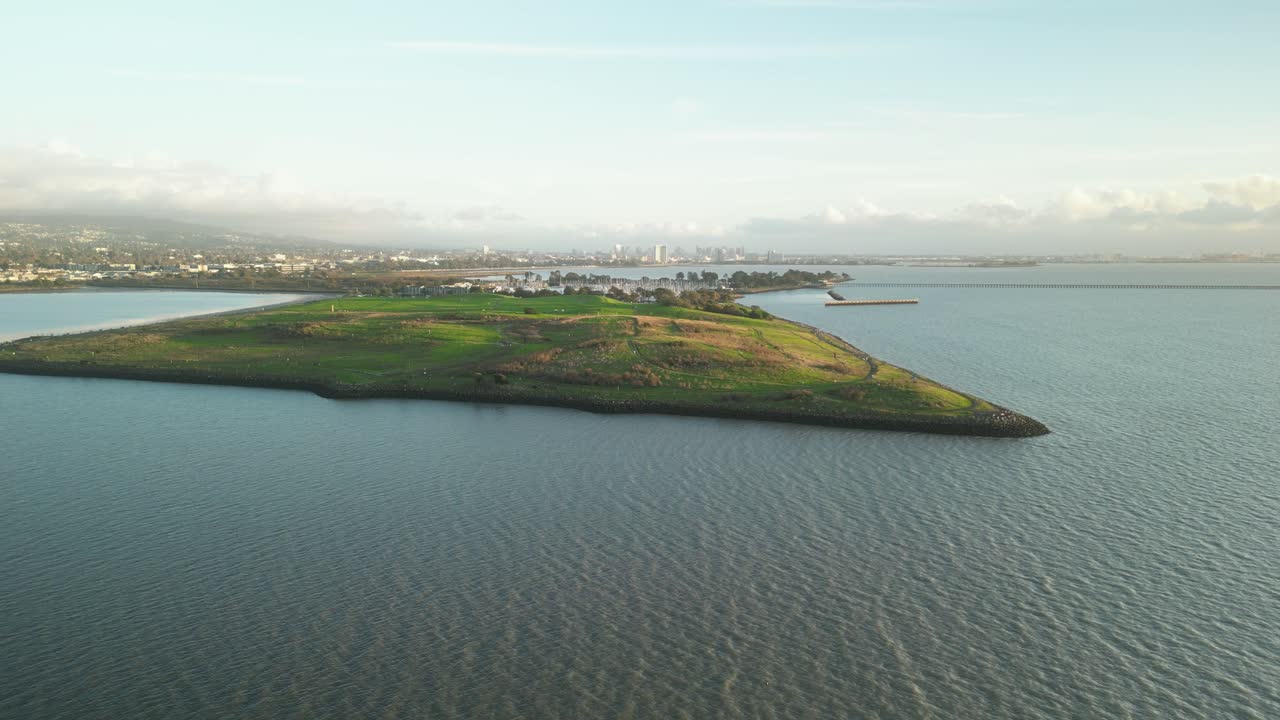 amplia vista aérea del parque césar chávez, una península que se extiende en la bahía de san francisco, con colinas cubiertas de hierba, aguas tranquilas y el paisaje urbano de berkeley en la distancia en un día parcialmente nublado