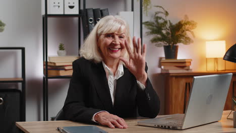 happy elderly businesswoman with laptop waves hand hi gesture greeting someone at home office desk