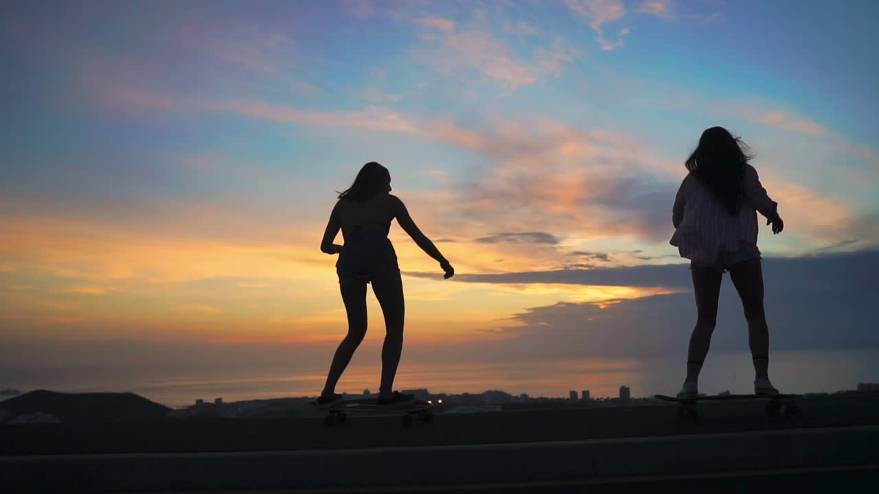 A sunset scene showcases two friends in slow motion, skateboarding on a road with mountains and a picturesque sky. They're dressed in shorts