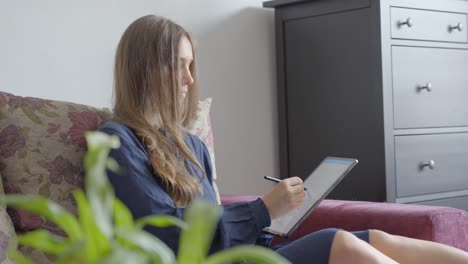 pretty young girl writing with stylus on tablet, view from side on sofa