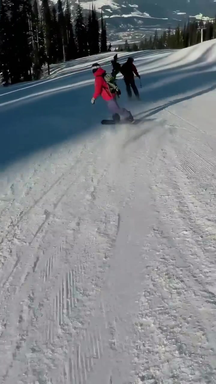 Video verticale: La telecamera segue uno snowboarder con una giacca rossa che scia su una pista battuta per divertimento, con uno zaino.