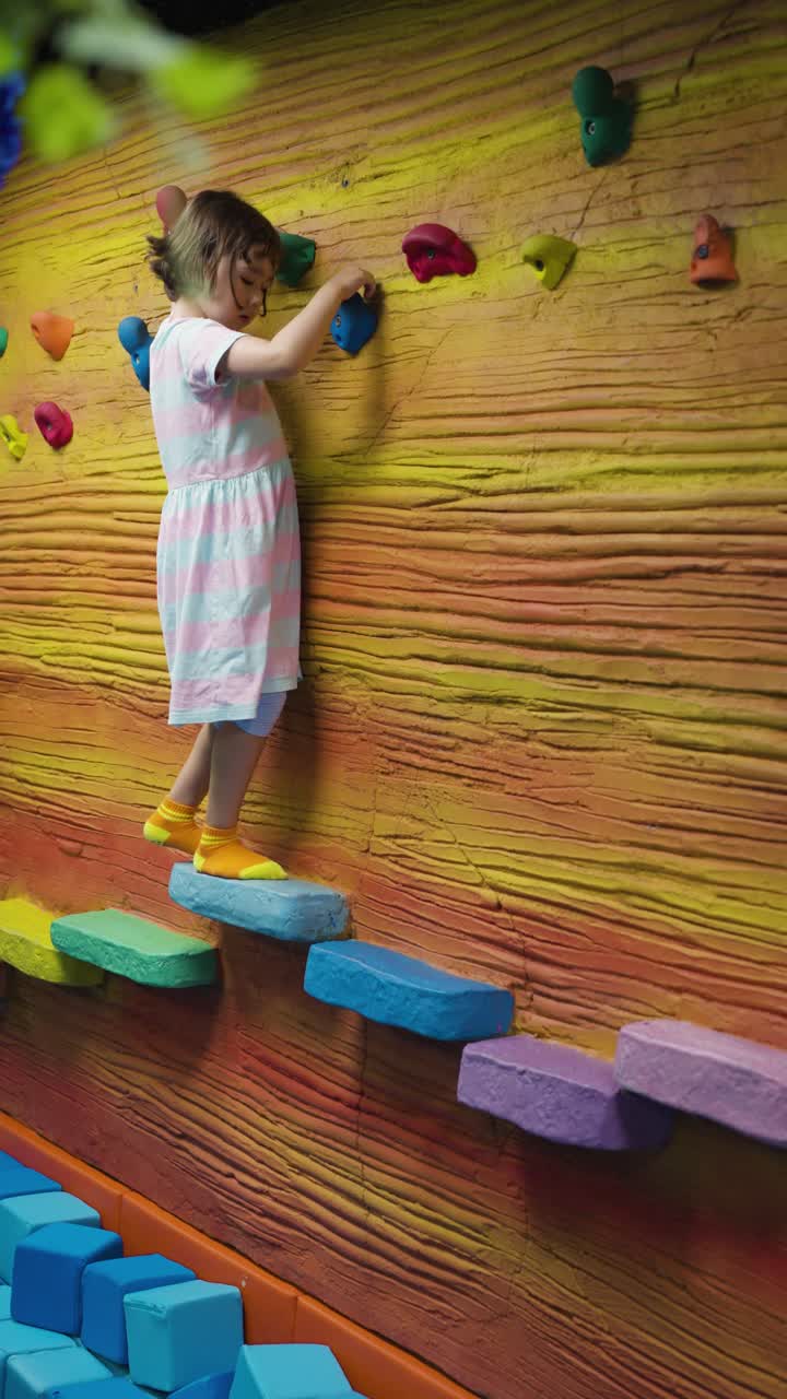 Vista lateral de una joven contenta y decidida que escala grandes apoyos para los pies, coloridos y con forma de escalera, en un centro de juegos interior