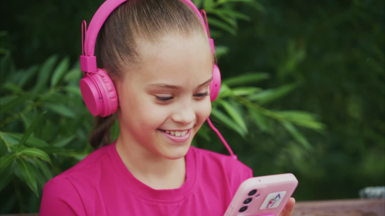 A Young Girl Enjoys Music and Games: Captured Moments of Joy with Pink Headphones and Smartphone in a Natural Setting