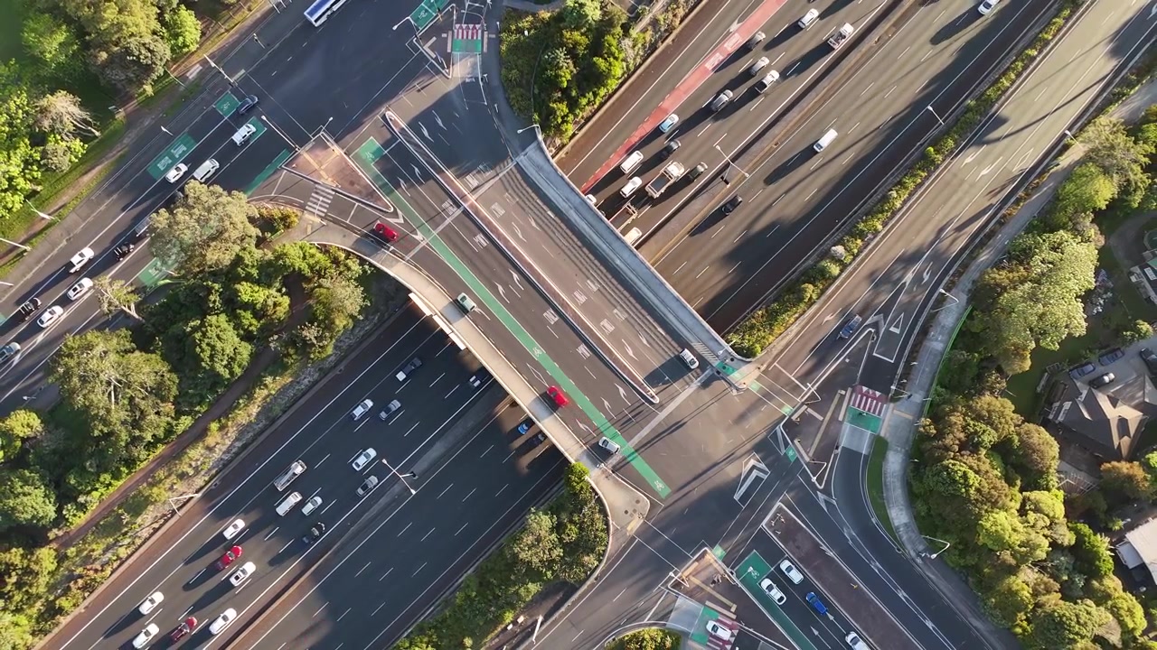 Overpass bridge carries traffic above busy Northwestern Motorway lanes in Auckland, New Zealand, cars merge through curved intersection as green cycle lane crosses bridge. Aerial drone top down shot