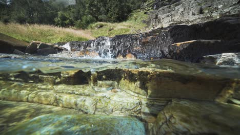 Bezaubernde-Über-Unter-Ansicht-Eines-Flachen-Flusses-Mit-Transparentem-Wasser-Und-Einem-Wasserfall