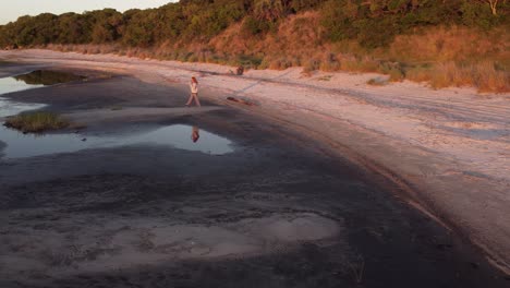 toma aérea de una mujer con el pelo rojo caminando por la orilla de la laguna al atardecer y haciendo una videollamada