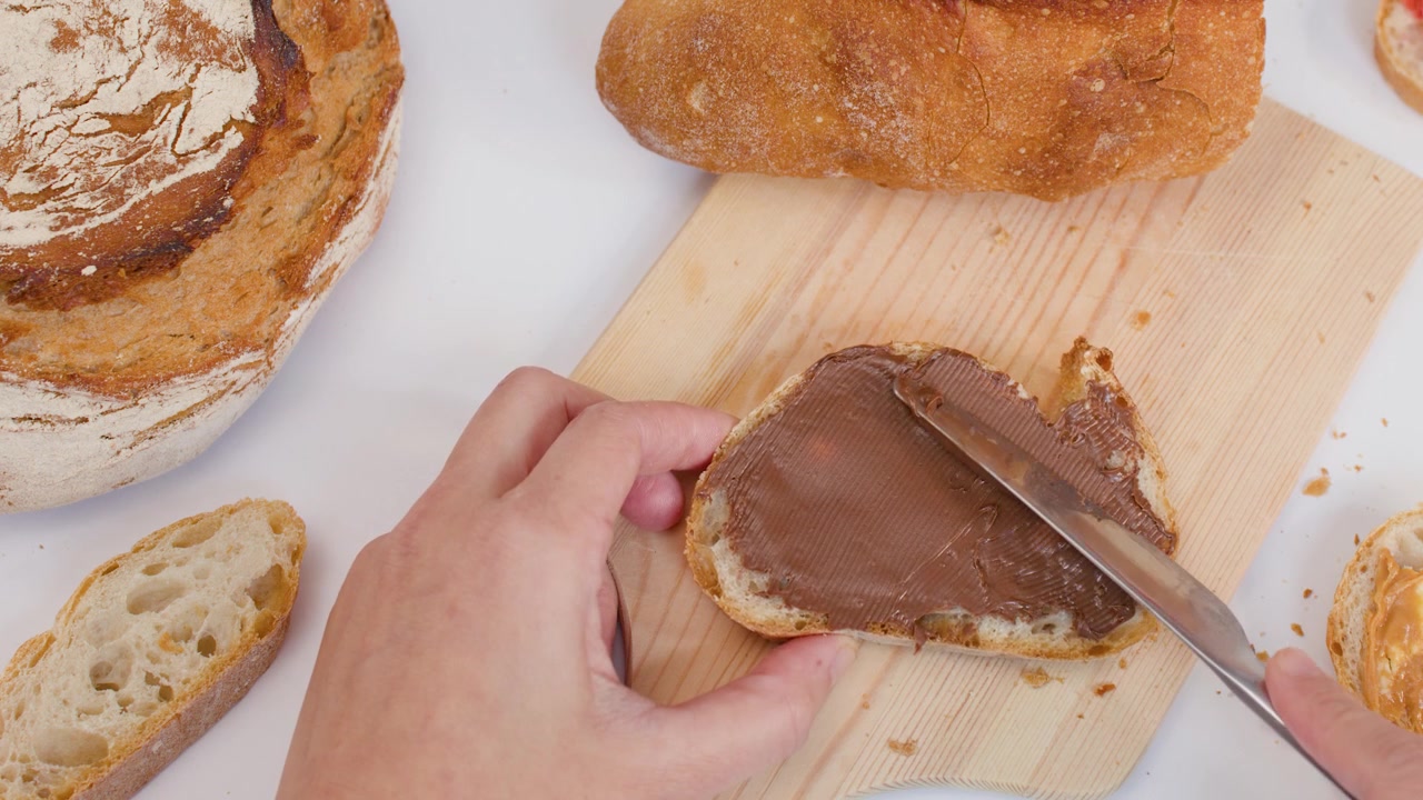 Vista dall'alto di una mano che usa un coltello per spalmare crema al cioccolato sul pane su un tagliere di legno.