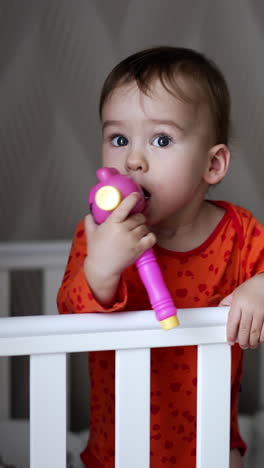 female hand gives a toy to a baby standing in the crib. sweet little baby takes a toy and shoves it to his mouth looking up. close up. vertical video