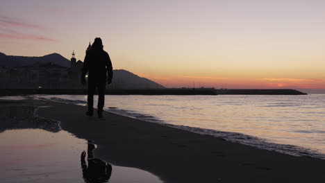 man walks along tranquil sea, dusky coastline stretches out before him, leading towards silhouette of a church set against the majestic mountain range