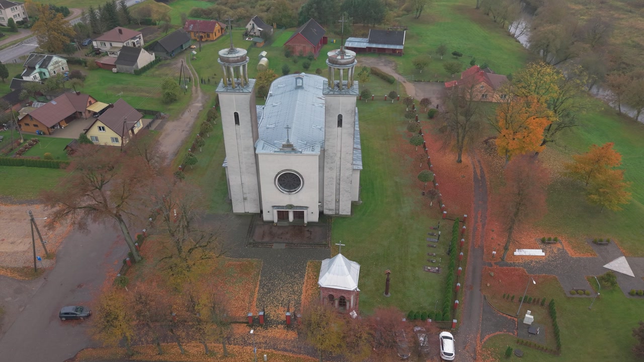 Drone footage captures Pilviškiai Holy Trinity Church draped in autumn hues. The scenic view showcases surrounding fields and architecture from a bird's eye perspective.