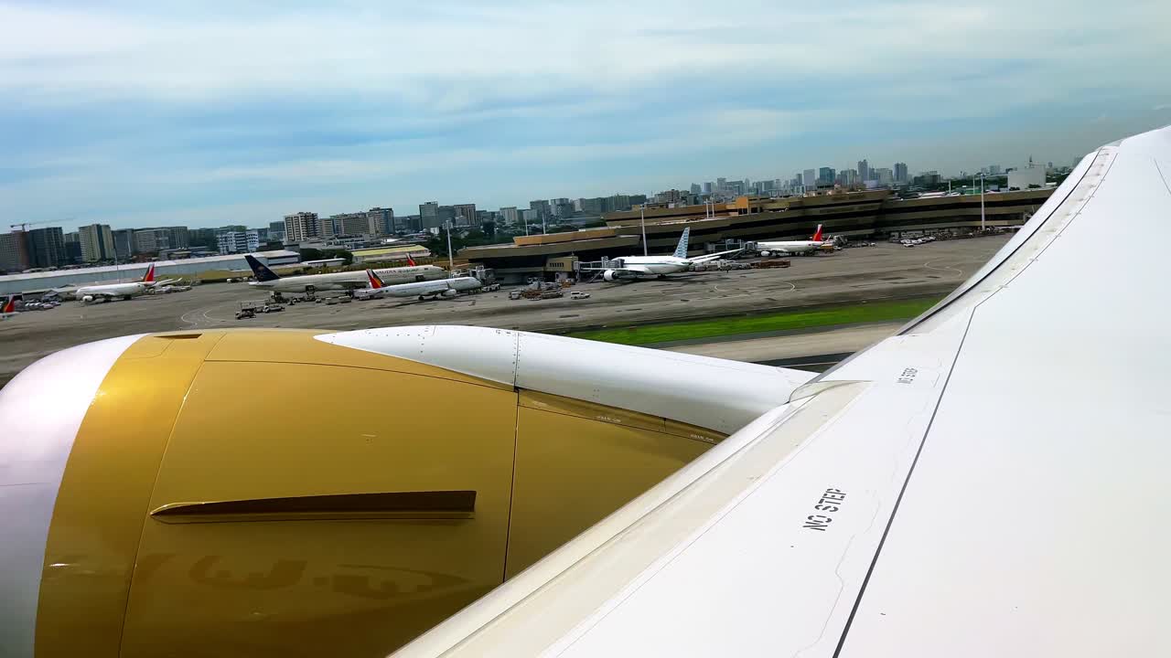 Close-up airplane window view of golden engine and wing positioned on runway, ready for takeoff, with airport buildings and control tower in the background