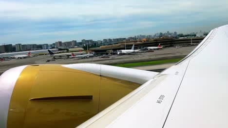 close-up airplane window view of golden engine and wing positioned on runway, ready for takeoff, with airport buildings and control tower in the background