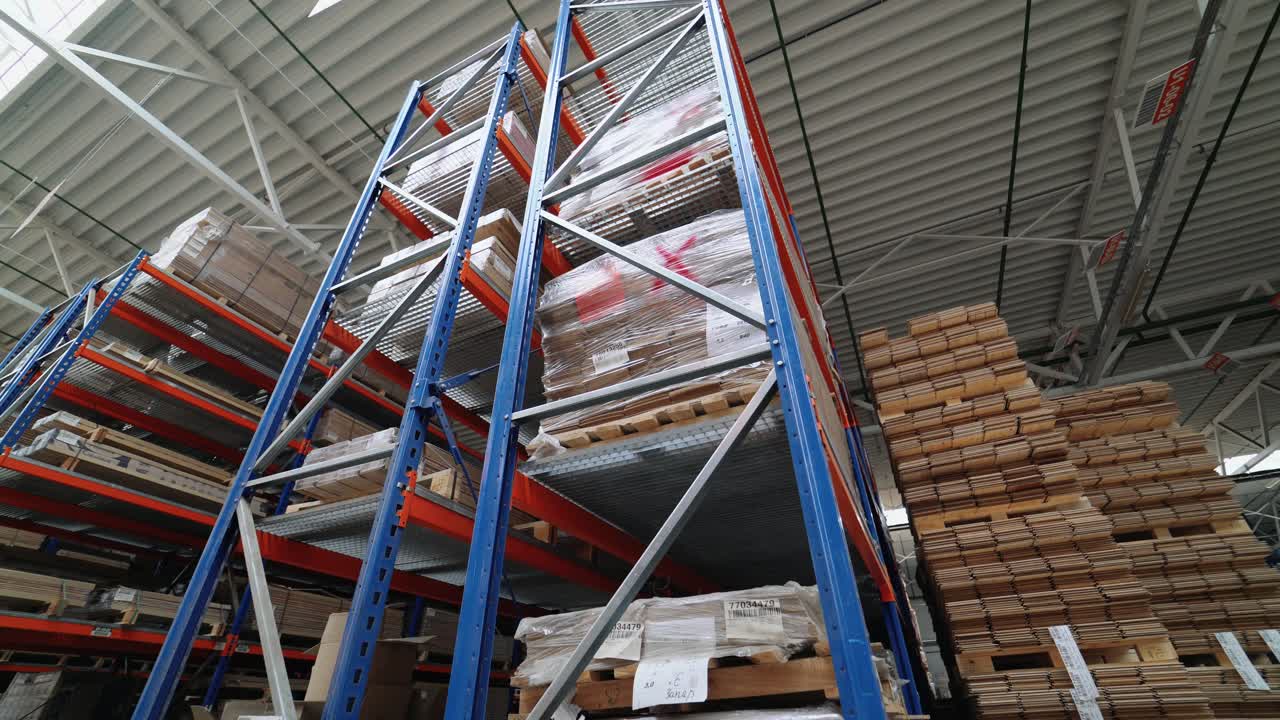 High metal shelves of blue color are filled with wooden boards in the building of a warehouse of a woodworking factory. View of the large storage of parquet from inside