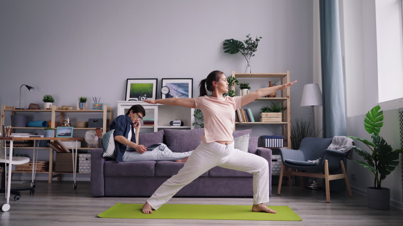 Woman practicing yoga at home while her partner relaxes on the couch