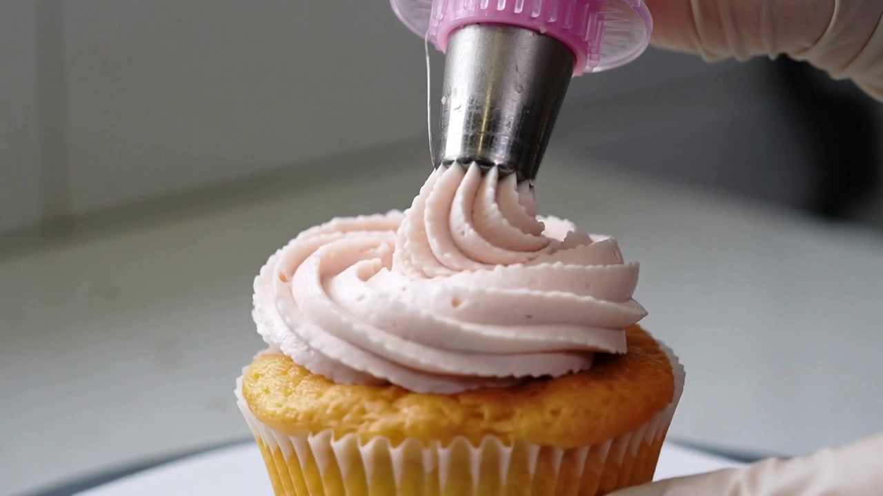 Tangan bersarung tangan memipet, memencet alat untuk menghias cupcake dengan hiasan putar merah muda pucat di konter, menggunakan spuit bintang.