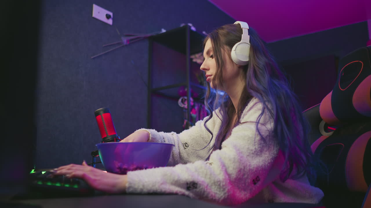 Young woman wearing headphones types on keyboard while eating popcorn from bowl, wipes hand on fluffy cardigan, then resumes typing in colorful neon lit workspace