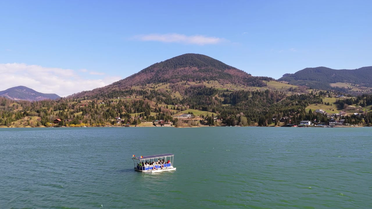 Vista aérea de drone de um barco navegando por águas cintilantes com montanhas em camadas ao fundo, no Lago Colibita, Romênia