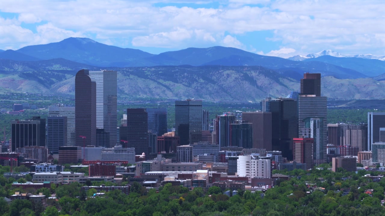 Drone-luftfoto af Denvers skyline med skyskrabere i downtown Denver, Colorado, mod et landskab med de sneklædte Rocky Mountains (Front Range), en forårsmorgen med blå, skyet himmel, filmet med en opadgående bevægelse baglæns