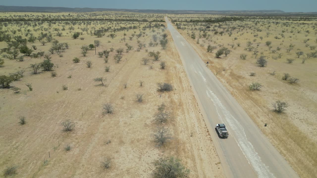 Vista aerea da drone nel deserto della Namibia, con un'auto SUV che percorre una strada stretta, sabbia e alberi verdi ai lati e montagne all'orizzonte in una giornata di sole